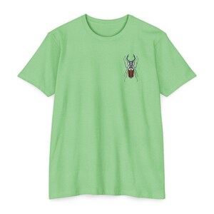 May include: Light green t-shirt with a detailed beetle illustration on the upper left chest. The beetle has a brown body, blue and white accents, and large, curved horns. The shirt has short sleeves and a crew neck.