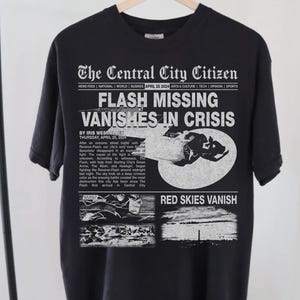 May include: Black t-shirt with a white print of a newspaper headline that reads "FLASH MISSING VANISHES IN CRISIS" with a comic book style image of a superhero in the background. The text "RED SKIES VANISH" is printed in the bottom right corner.