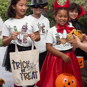 May include: Four children dressed in Halloween costumes, holding trick-or-treat bags and candy. The girl on the left is wearing a white shirt with the word "LOVE" in colorful letters, a witch hat, and a striped skirt. The boy in the middle is wearing a black cowboy hat, a black mask, and a white shirt with the words "For the LOVE Halloween". The girl on the right is wearing a red devil costume with horns and a red skirt. The boy on the far right is wearing a vampire costume with a black cape. The girl on the left is holding a white canvas tote bag with the words "Trick or Treat" in colorful letters.