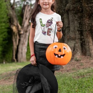 May include: A young person dressed as a witch for Halloween, holding a bright orange pumpkin bucket with a smiling face. They are wearing a white shirt with the word "LOVE" spelled out in colorful, Halloween-themed letters, a black skirt, and a black witch hat with a veil.