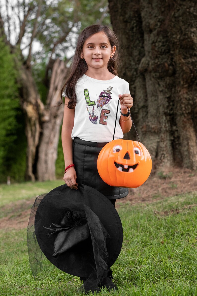 May include: A young person dressed as a witch for Halloween, holding a bright orange pumpkin bucket with a smiling face. They are wearing a white shirt with the word "LOVE" spelled out in colorful, Halloween-themed letters, a black skirt, and a black witch hat with a veil.