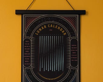 May include: A black lunar calendar print with a decorative border and the words "Lunar Calendar 2026." The calendar shows moon phases and inspirational quotes. The print is held by black wooden bars and hangs on a yellow wall.