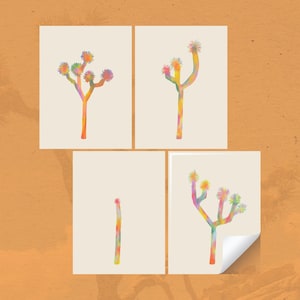 Könnte beinhalten: Vier Kunstdrucke mit Aquarell-Illustrationen von Joshua-Bäumen. Jeder Baum ist in leuchtenden Regenbogenfarben auf cremefarbenem Hintergrund dargestellt. Die Drucke sind auf einem strukturierten, orangefarbenen Hintergrund angeordnet, mit einer umgeknickten Ecke.