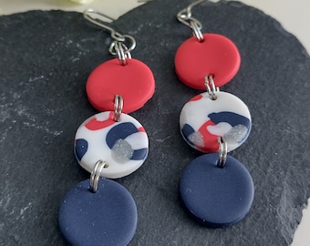 May include: A pair of dangle earrings with three round polymer clay beads. The top bead is red, the middle bead is white with black and red spots, and the bottom bead is navy blue.