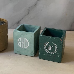 May include: Two small, square, green concrete planters with white lettering. One planter has the initials "GHD" and the other has the letter "F" inside a wreath.