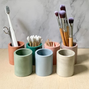 Puede incluir: Seis pequeños contenedores de hormigón de varios colores, incluyendo verde, azul y rosa, están dispuestos en una fila. Los contenedores están llenos de varios artículos, incluyendo bastoncillos de algodón, palillos de dientes y pinceles de maquillaje.