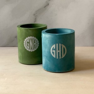 Può includere: Due vasi cilindrici in cemento, uno verde e uno blu, con iniziali monogramma bianche. Il vaso verde ha le iniziali "GNB" e il vaso blu ha le iniziali "GHD".