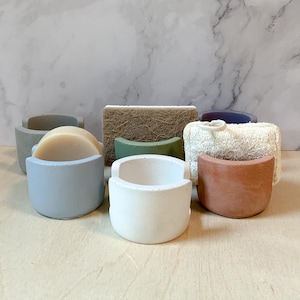 May include: Six soap dishes made of concrete in various colors, including light blue, white, and terracotta. The dishes are designed to hold soap bars and feature a curved shape. A bar of soap is sitting in one of the dishes.