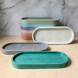 Puede incluir: Un conjunto de bandejas ovaladas de hormigón en varios colores, incluyendo verde, gris, blanco, rosa, azul y beige. Las bandejas están apiladas unas sobre otras, con la bandeja verde en la parte delantera.
