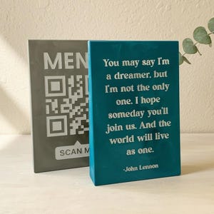 May include: A rectangular teal sign featuring white text that reads, "You may say I'm a dreamer, but I'm not the only one. I hope someday you'll join us. And the world will live as one. -John Lennon." Behind it stands a grey sign with a QR code.