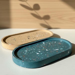 Könnte beinhalten: Zwei ovale Tabletts mit Terrazzo-Optik. Ein Tablett ist in einem tiefen Blaugrün mit bunten Sprenkeln gehalten, das andere in einem hellen Beige mit ähnlichen Sprenkeln. Beide Tabletts haben eine glatte, flache Oberfläche mit erhöhtem Rand.