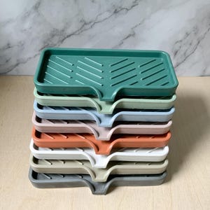 May include: A stack of nine rectangular dish drying racks in various colors, including green, blue, pink, orange, white, and gray. The racks have a raised, textured surface and a curved edge.