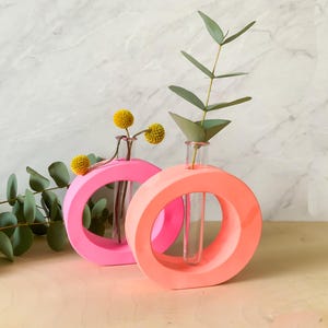 Puede incluir: Dos jarrones modernos y minimalistas en rosa brillante y coral. Cada jarrón tiene un diseño circular con un tubo de vidrio transparente en el centro, que contiene flores amarillas y eucalipto. Los jarrones están sobre una superficie de madera clara, con fondo de mármol.