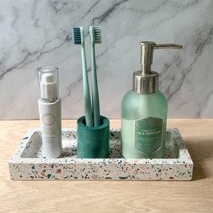 Puede incluir: Una bandeja de terrazo blanco con un soporte de cerámica verde para dos cepillos de dientes, una botella de jabón de manos etiquetada como "Capri Blue Fir & Firewood", y una botella de tratamiento purificante para imperfecciones.