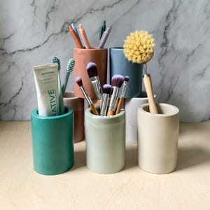 Puede incluir: Cinco contenedores cilíndricos de hormigón en varios colores, incluyendo verde, beige y verde claro, que contienen varios artículos como cepillos de dientes, pinceles de maquillaje y un cepillo de limpieza. Los contenedores están sobre una superficie de madera clara.