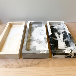 May include: Three rectangular concrete trays. The tray on the left is a plain white color. The middle tray is a light gray with white marbling. The tray on the right is black and white with marbling.