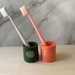 Puede incluir: Dos portacepillos de dientes, uno verde y otro coral, con monogramas personalizados. El portacepillos verde tiene el monograma "GHD" y el portacepillos coral tiene el monograma "JID".