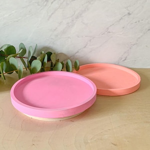 May include: Two round decorative trays. One is a bright pink, the other is a soft coral color. Both have a raised edge and sit on a light wood base. The trays are empty and ready for use.