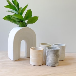 May include: Four small white concrete planters with a speckled finish. The planters are arranged in a row on a wooden surface. A larger white concrete planter shaped like an arch is in the background.