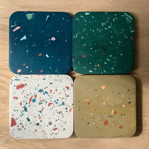 May include: Four square coasters with a terrazzo design. The coasters are made of a light gray material with flecks of green, red, and blue. The coasters are arranged in a square pattern.