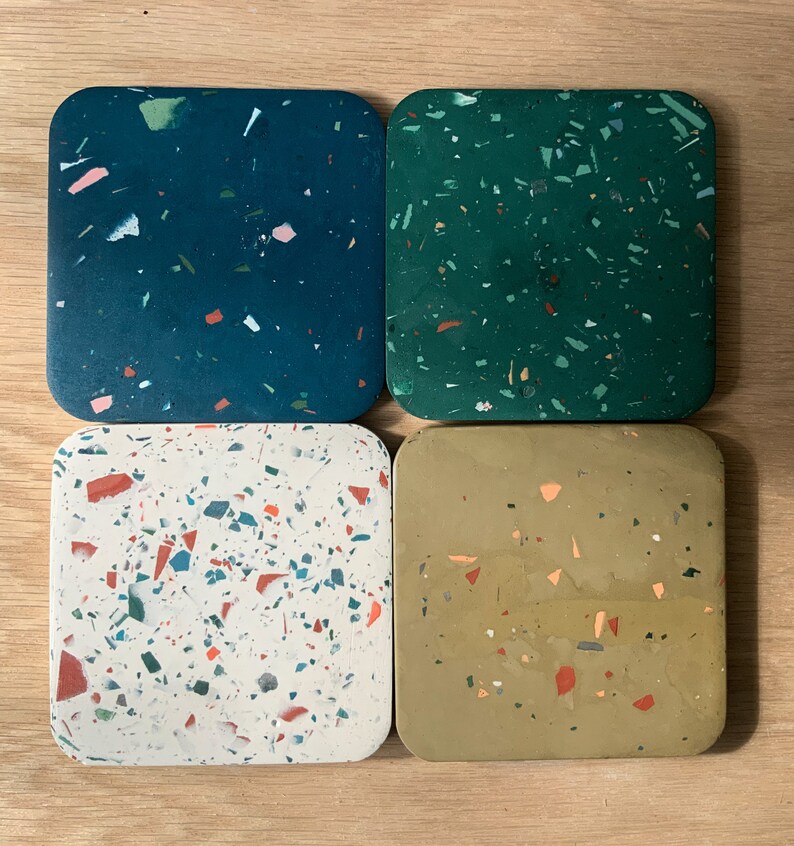 May include: Four square coasters with a terrazzo design. The coasters are made of a light gray material with flecks of green, red, and blue. The coasters are arranged in a square pattern.