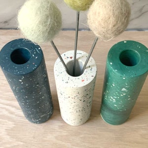 May include: Three cylindrical vases with speckled patterns in black, white, and green. The vases are filled with three faux flower stems with round, fluffy balls in yellow, white, and green.