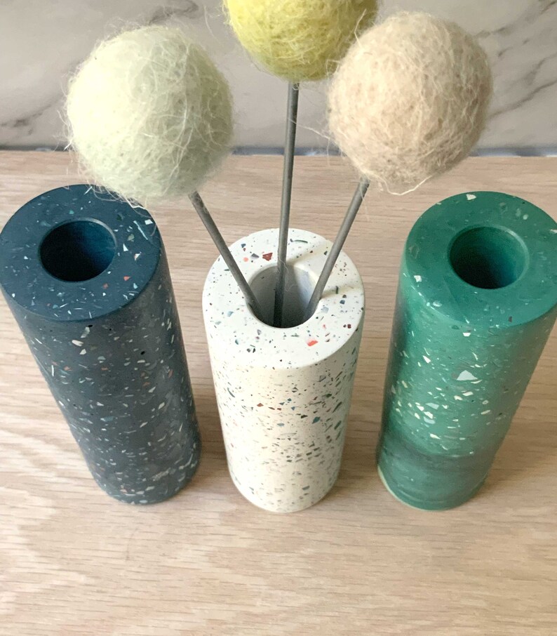 May include: Three cylindrical vases with speckled patterns in black, white, and green. The vases are filled with three faux flower stems with round, fluffy balls in yellow, white, and green.