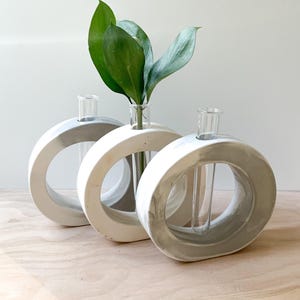 Puede incluir: Tres jarrones de cerámica blanca y gris con aberturas redondas y tubos de ensayo de vidrio transparente para colocar flores. Los jarrones están dispuestos en fila sobre una superficie de madera clara.