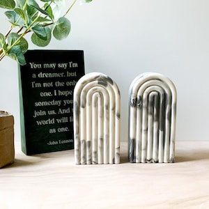 May include: Two white and gray marble-look bookends shaped like arches with rounded tops. The bookends have a grooved design and are a modern home decor accent.