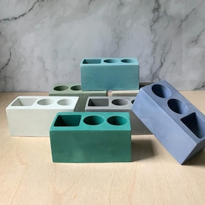 May include: Six concrete planters in various colors, each with three round holes for planting. The planters are shaped like bricks and are arranged in a staggered pattern.