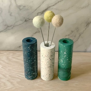 May include: Three cylindrical vases made of speckled terrazzo in blue, white, and green. The vases are arranged in a row on a wooden surface. Three small felt balls in yellow, light green, and beige are placed in the white vase.