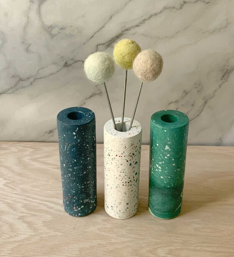 May include: Three cylindrical vases made of speckled terrazzo in blue, white, and green. The vases are arranged in a row on a wooden surface. Three small felt balls in yellow, light green, and beige are placed in the white vase.