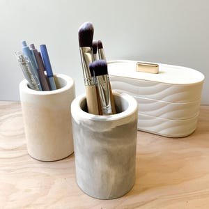 May include: Two white ceramic cylindrical containers, one with a marble pattern, holding makeup brushes and pens. A white oval container with a gold top is in the background.