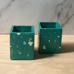 Peut inclure: Deux pots carrés en terrazzo de couleur verte avec des taches blanches et brunes. Les pots sont en béton et ont une finition lisse.