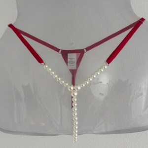 May include: A red thong featuring thin adjustable straps and a central vertical strand of white faux pearls. The garment includes a small white care label attached to the center back, with a minimalist design that highlights the contrast between the vibrant red fabric and the smooth, round white pearls.