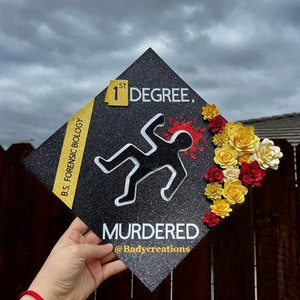Op de afbeelding: Een zwarte afstudeerhoed met glitter en een witte omtrek van een persoon die op de grond ligt met een rode bloedvlek. De hoed heeft een geel lint met de tekst "B.S. FORENSIC BIOLOGY" en een geel vierkant met de tekst "1ST DEGREE." De tekst "MURDERED" staat in wit onder de omtrek van de persoon. De tekst "@Badycreations" staat in wit onder de tekst "MURDERED." De hoed is versierd met rode, gele en witte papieren bloemen.