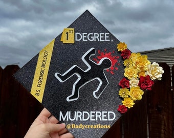 Puede incluir: Un gorro de graduación negro con brillantina y un contorno blanco de una persona tendida en el suelo con una salpicadura de sangre roja. El gorro tiene una cinta amarilla con el texto "B.S. FORENSIC BIOLOGY" y un cuadrado amarillo con el texto "1ST DEGREE." El texto "MURDERED" está escrito en blanco debajo del contorno de la persona. El texto "@Badycreations" está escrito en blanco debajo del texto "MURDERED." El gorro está decorado con flores de papel rojas, amarillas y blancas.