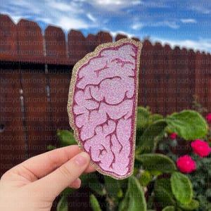 Könnte beinhalten: Eine rosa glitzernde, gehirnförmige Dekoration mit goldenem Rand. Das Gehirndesign weist detaillierte Rippen und eine glitzernde Oberfläche auf. Die Dekoration wird vor einem Hintergrund aus grünem Laub und einem Holzzaun gehalten.