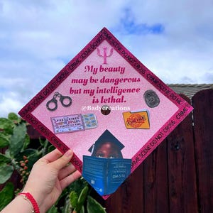 Op de afbeelding: Een roze glitter afstudeerhoed met de tekst "Mijn schoonheid kan gevaarlijk zijn, maar mijn intelligentie is dodelijk." Voorzien van handboeien, een vingerafdruk en een boek. De rand luidt "CRIME SCENE DO NOT CROSS."