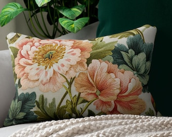 May include: A decorative rectangular pillow with a floral design featuring large pink and peach flowers with green leaves. The pillow is on a gray sofa with a green pillow and a cream-colored blanket.