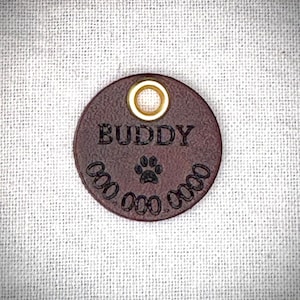 May include: A round, dark brown leather pet tag with the name "BUDDY" embossed in capital letters. A paw print is centered below the name, and a phone number is below the paw print. The tag has a gold-colored metal eyelet.