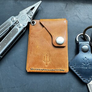 May include: A brown leather wallet with a stitched edge and a silver button. The wallet has a trident logo embossed on the front.