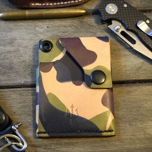 May include: A camouflage leather wallet with a black snap closure and a trident logo. The wallet is made of high-quality leather and has a unique design.