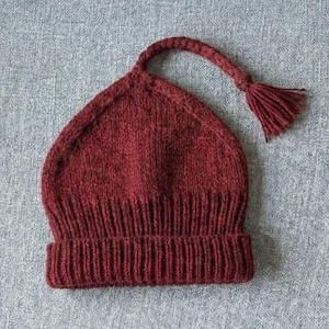 Pode incluir: Um gorro bordô de malha com uma aba dobrada e uma longa alça trançada que termina em uma borla. O chapéu é feito de um fio texturizado e fica em uma superfície cinza. O design do chapéu é simples e clássico.