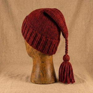 Op de afbeelding: Een bordeauxrode gebreide muts met een lange, puntige bovenkant en een kwast. De muts wordt getoond op een houten hoofd tegen een neutrale achtergrond. De textuur en kleur van de muts zijn prominent aanwezig.