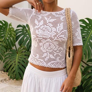 May include: A white short-sleeved crochet top featuring an intricate floral pattern throughout the open-knit fabric. The garment has a round neckline and a straight hemline. A tan woven straw tote bag with braided handles is carried on the shoulder.