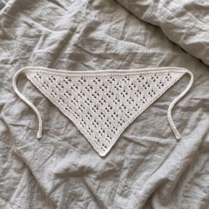 Könnte beinhalten: Ein weißes, dreieckiges Strickbandana mit einem filigranen Lochmuster. Das Bandana hat lange, dünne Bindebänder an jeder Ecke zum Befestigen. Der Stoff wirkt weich und leicht, geeignet für warmes Wetter.