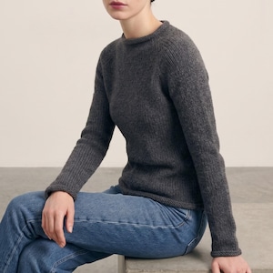 Peut inclure: Un pull en tricot gris anthracite à manches longues avec une texture côtelée et un col rond, associé à un jean bleu. Le pull présente des manches raglan et une silhouette simple et ajustée qui arrive aux hanches, offrant un style classique.