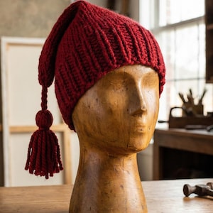 Op de afbeelding: Een gebreide, dieprode muts met een lange, puntige punt en een kwast, tentoongesteld op een houten mannequinhoofd. De geribbelde textuur en rijke kleur van de muts zijn zichtbaar. De achtergrond omvat een houten tafel en een raam.