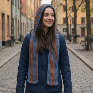 May include: A blue and orange striped hooded scarf is worn over a dark blue jacket. The scarf is made of a knitted material and has a long, rectangular shape. The person is smiling and walking on a cobblestone street.